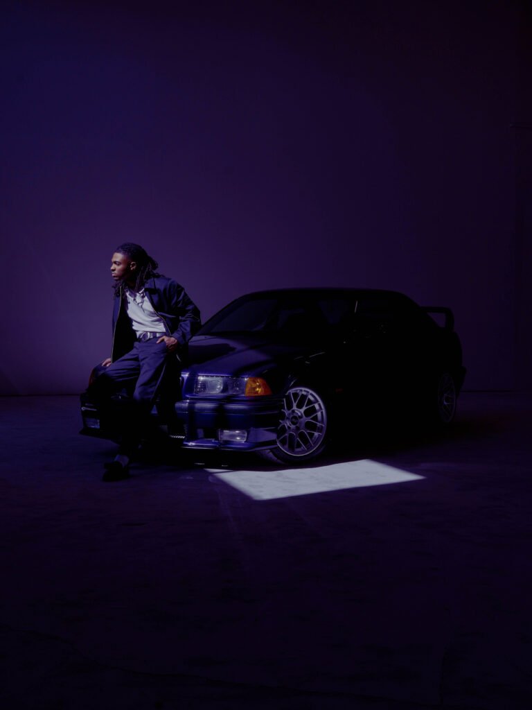 A photo of singer Tone Stith leaning against a car, looking away from the camera